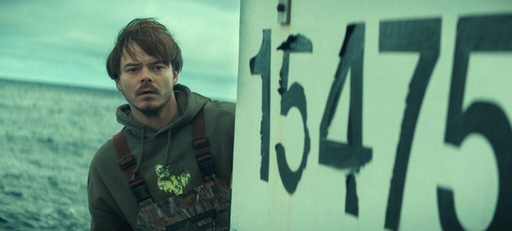 "A man in fishing gear stands on a boat at sea, with a large boat number visible, under a cloudy sky."
